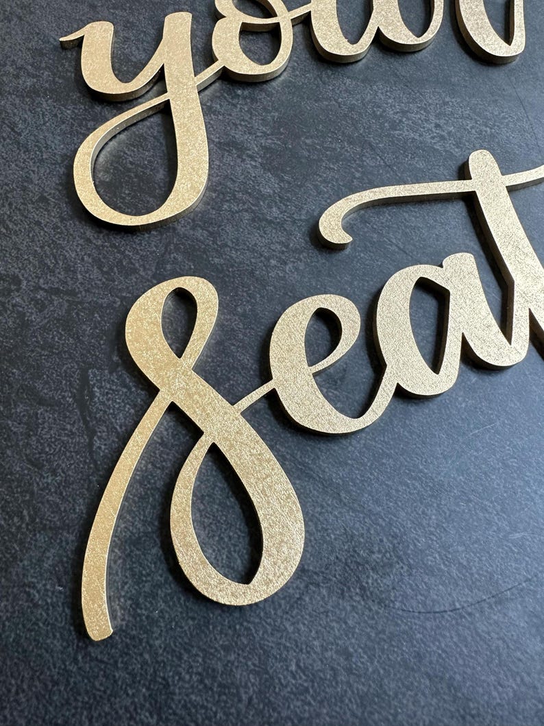 May include: Gold-colored script lettering cutouts spelling the words "your seat". The letters have a textured, metallic finish and are presented in a cursive font style with connected characters, creating a decorative sign for event seating.