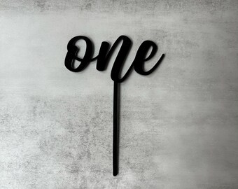 May include: A black cake topper featuring the word one in a cursive script font. The design consists of the lowercase letters o, n, and e connected together, attached to a single vertical stake extending downward from the base of the lettering.