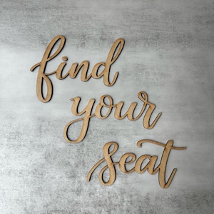 May include: A set of three wooden cutout words in a cursive script font that spell out the phrase find your seat. The letters have a natural light brown wood finish with visible grain and are arranged in three separate lines on a gray surface.