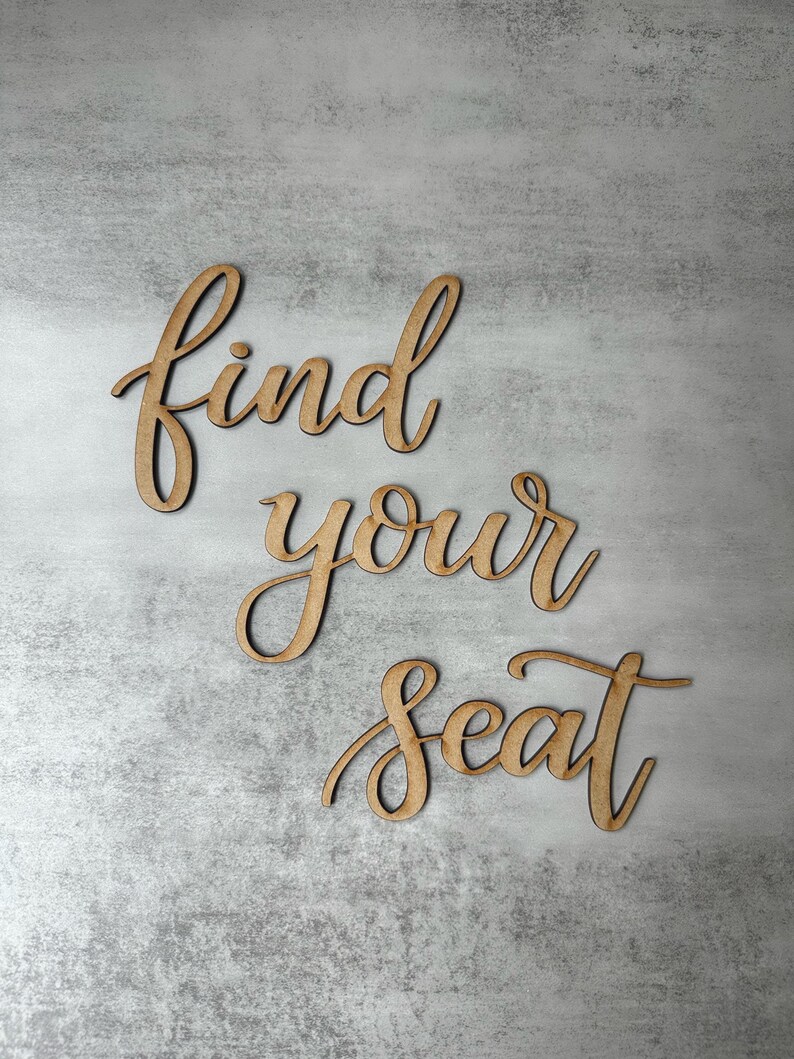 May include: A set of three wooden cutout words in a cursive script font that spell out the phrase find your seat. The letters have a natural light brown wood finish with visible grain and are arranged in three separate lines on a gray surface.