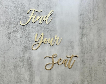 May include: Gold-colored script lettering spelling out the phrase Find Your Seat. The letters are individual cutouts with a metallic finish, arranged in three lines to form the complete message against a light gray textured background.