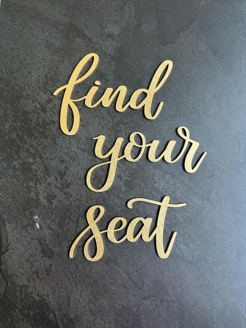 May include: Gold-colored script lettering cutouts spelling out the phrase find your seat. The letters feature a textured, metallic finish and are arranged in three separate lines, with each word positioned vertically below the previous one.
