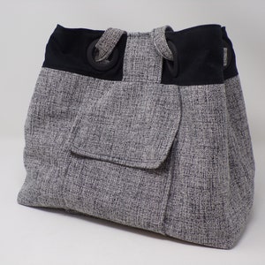 Può includere: Una borsa tote grigia e nera con una grande tasca sul davanti. La borsa ha manici neri e anelli neri sui manici.