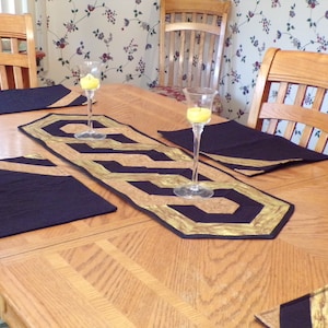Peut inclure: Un chemin de table noir et or avec un motif géométrique, placé sur une table en bois avec des sets de table noirs. Deux bougeoirs en verre transparent avec des bougies jaunes sont sur la table.