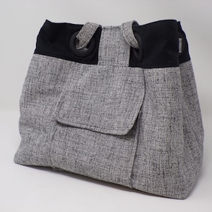 Può includere: Una borsa tote grigia e nera con una grande tasca sul davanti. La borsa ha manici neri e anelli neri sui manici.