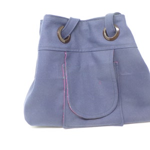 Può includere: Una borsa tote in tela blu scuro con due anelli di metallo nero in cima e una grande tasca sul davanti. La tasca ha un bordo rosa.