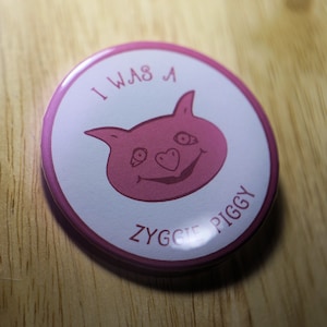 Puede incluir: Un botón rosa con el texto "I was a Zyggie Piggy" y un dibujo animado de un cerdo con una nariz en forma de corazón.