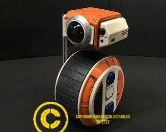 以下が含まれることがあります： 白、オレンジ、黒のロボットで、車輪が1つあります。ロボットにはアンテナが2本と丸いカメラレンズが付いています。ロボットは黒い背景の上にあります。Copyright BigBangCollectibles on Etsy.
