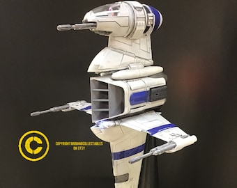 May include: A white and blue Star Wars V-4X-D ski speeder model with a black stand. The model has a detailed cockpit and wings with blue and white stripes. The model is on a black stand with a round base.