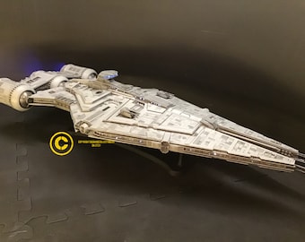 May include: A white model of a spaceship with a long, narrow body and a pointed nose. The ship has a large, flat wing on each side and a smaller wing on the top. The ship is sitting on a black surface.
