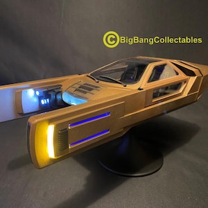 Puede incluir: Modelo dorado de un vehículo futurista con luces azules y amarillas. El vehículo tiene un diseño elegante con una cabina transparente y está montado sobre una base negra. El logotipo de BigBangCollectables es visible.