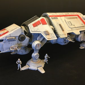 以下が含まれることがあります： 白と赤のディテールが施されたスターウォーズAT-TEウォーカーのモデル。その前に白いフィギュアが3体立っています。