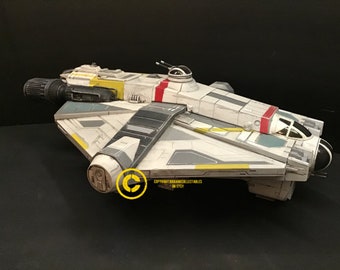 May include: A white and gray spaceship toy with yellow and red accents. The spaceship has a long, cylindrical engine and a cockpit with a clear canopy. The toy is made of plastic and has a detailed design.
