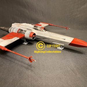 Può includere: Un modello rosso, bianco e grigio di un caccia X-wing di Star Wars. Il modello è dettagliato con una cabina di pilotaggio, motori e ali. Il modello è seduto su una superficie grigia. COPYRIGHT BigBangCollectibles