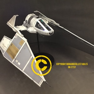 May include: A white model of a Star Wars X-wing fighter with black wings and a gray cockpit. The model is on a black stand with a copyright symbol and the text "COPYRIGHT BIGBANGCOLLECTABLES ON ETSY".