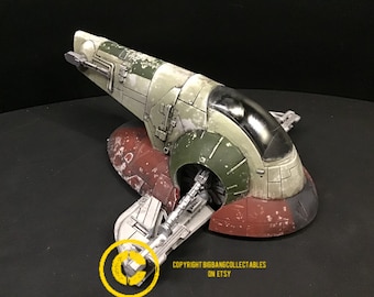 May include: A green and brown model of a Star Wars Slave I spaceship. The model is weathered and has a worn paint finish. The model is on a black surface.