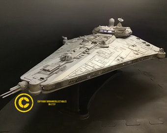 May include: A detailed model of a Star Destroyer spaceship, mainly grey with intricate paneling and detailing. The model features multiple turrets, a central command structure, and a radar dish. The ship is mounted on a clear stand against a dark background.