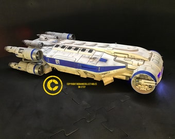 May include: A white and blue model spaceship with detailed engine and cockpit features. The model has blue accents and lights. The model is on a black surface.