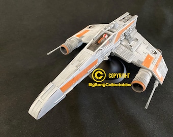 May include: A white and orange X-wing starfighter model with a pilot figure in the cockpit. The model is on a black base with the text "COPYRIGHT BigBangCollectables".