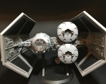 May include: A detailed model of a TIE fighter spacecraft, with a central cockpit sphere, two side-mounted spherical engines, and hexagonal solar panels. The model is grey and black, mounted on a black stand.
