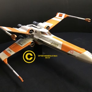 May include: A white and orange X-Wing starfighter model with the number "V-85" printed on the side. The model is on a black surface.
