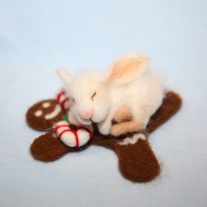 May include: A white felted bunny with pink ears and nose, sleeping on a brown felt gingerbread man. A red and white candy cane is next to the bunny. The background is a light blue.