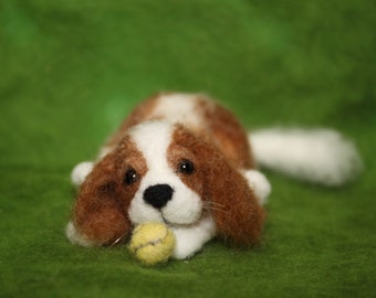Puede incluir: Una figura de perro de lana fieltrada con pelaje marrón y blanco, orejas largas y caídas y ojos oscuros, tumbada en posición de descanso. El perro sostiene entre sus patas delanteras una pequeña pelota amarilla texturizada con una costura gris.
