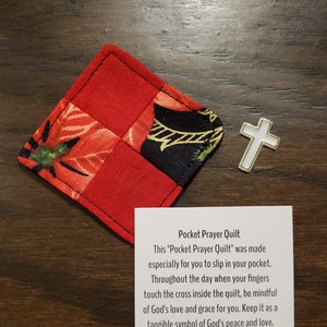 May include: A red and patterned pocket prayer quilt with a white cross. The quilt is made of squares of red fabric and fabric with floral and geometric patterns. A card reads "Pocket Prayer Quilt".