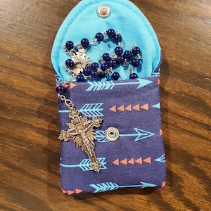 Puede incluir: Una bolsa de tela azul y azul marino con cierre de cremallera y un patrón de flechas turquesa y naranja. La bolsa está abierta y contiene un rosario plateado con cuentas azul oscuro.
