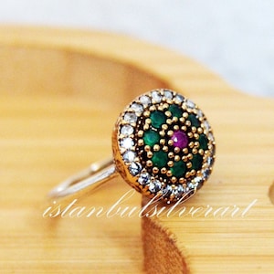 May include: A silver ring featuring a round green and pink gemstone encircled by smaller clear stones.