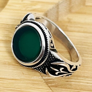 May include: A silver ring with a green stone set in a circular bezel. The ring has an ornate design on the band.
