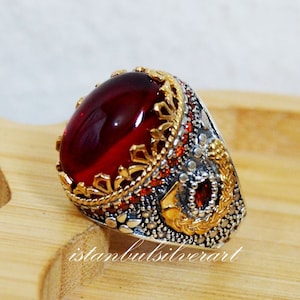 May include: A silver ring featuring a large red gemstone set within a gold crown-like design. The ring is adorned with smaller red gemstones and intricate detailing.