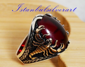 Puede incluir: Un anillo de plata con una piedra roja y un diseño de escorpión. El anillo está colocado sobre una superficie de madera.