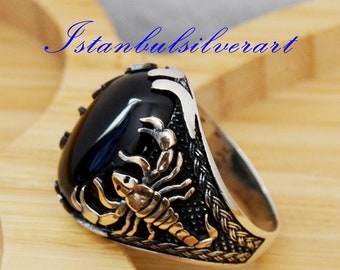 Puede incluir: Un anillo de plata con una piedra de ónix negro y un diseño de escorpión detallado. El anillo está sobre una superficie de madera.