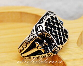 May include: A silver ring with a detailed design featuring a sword and a shield. The shield has a black and white checkered pattern.