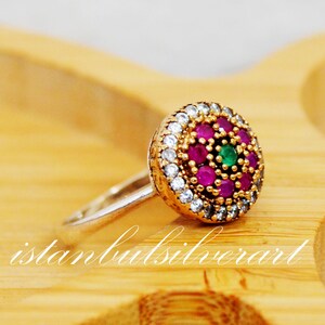 May include: A silver ring with a round, multi-colored gemstone setting. The ring features a cluster of pink and green stones surrounded by smaller white stones.