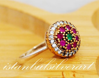 Può includere: Un anello d'argento con una montatura rotonda di pietre multicolori. L'anello presenta un gruppo di pietre rosa e verdi circondate da pietre bianche più piccole.