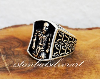 May include: A silver ring with a black and gold design featuring a warrior figure. The ring is on a wooden surface.