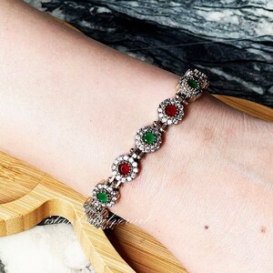May include: A silver bracelet with alternating red and green gemstones set in a floral design.