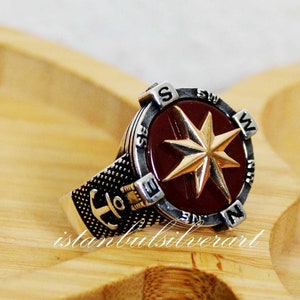 May include: A silver ring with a compass design. The compass face is red with a gold star and the compass points are marked with the letters N, S, E, and W. The ring band is textured and has an anchor design.