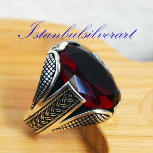Puede incluir: Un anillo de plata con una gran piedra roja de forma ovalada. El anillo tiene detalles intrincados y un diseño trenzado.