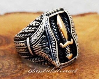 May include: A silver ring with a detailed design featuring a gold dagger on a black shield. The ring has a braided band.