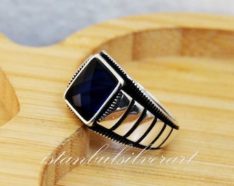 May include: A silver ring with a dark blue square-shaped gemstone. The ring has a textured band with multiple lines.