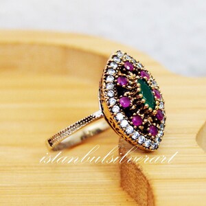 May include: A silver ring with a green and pink gemstone design. The ring is set on a wooden surface.