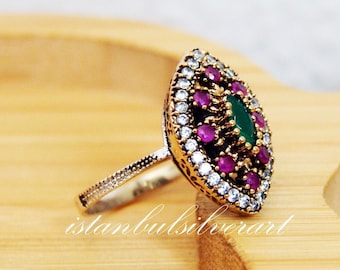 Può includere: Un anello d'argento con un designo di pietre preziose verdi e rosa. L'anello è posto su una superficie di legno.