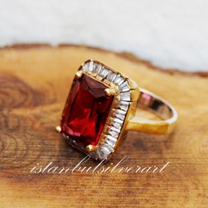 Puede incluir: Un anillo de oro con una gran piedra rectangular roja rodeada de piedras más pequeñas y transparentes. El anillo está sobre una superficie de madera.