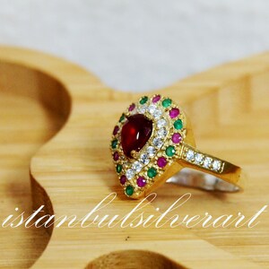 May include: A gold ring with a red heart-shaped stone surrounded by smaller white and green stones.