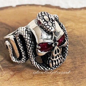 Peut inclure: Bague en argent en forme de crâne avec des yeux en pierres précieuses rouges et un serpent enroulé autour du crâne. Le serpent a une pierre précieuse rouge sur la tête.