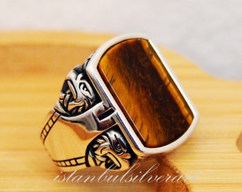 May include: A silver ring with a large, rectangular tiger's eye stone set in a detailed, ornate design. The ring has a textured band with intricate details.