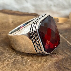 May include: A silver ring with a red gemstone set in a square bezel. The ring has a decorative border around the stone.